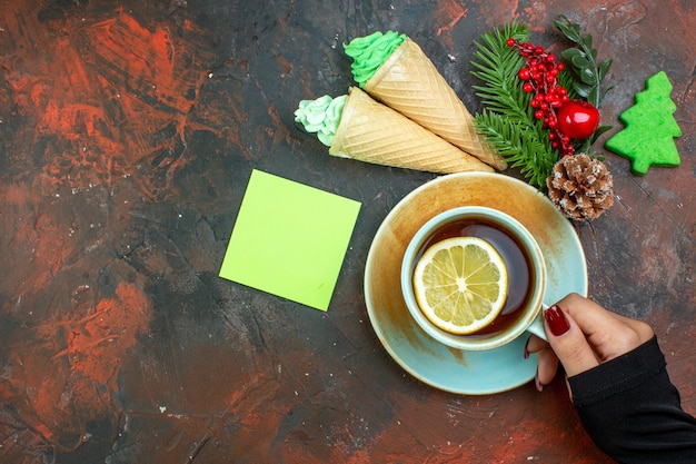 Free photo top view cup of tea flavored by lemon in female hand xmas tree branch ice creams sticky note on dark red table with free place