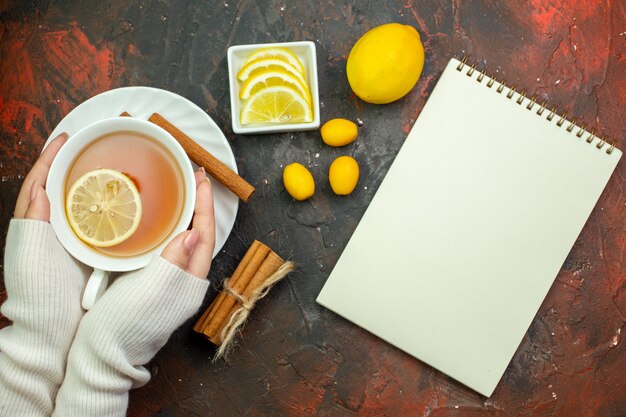 Top view cup of tea flavored by lemon in woman hands cumcuat lemon slices in small bowl tied cinnamon sticks notebook on dark red table