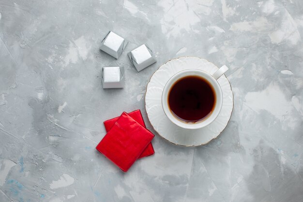 Top view of cup of tea hot inside white cup on glass plate with silver package chocolate candies on light desk, tea drink sweet chocolate