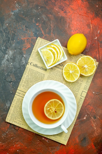 Top view cup of tea lemon slices in small bowl on newspaper on dark red background