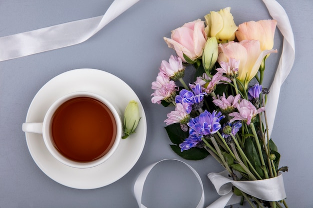 Top view of cup of tea on saucer and flowers with ribbon on gray background