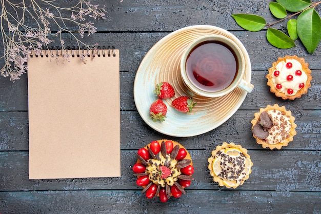 Free photo top view a cup of tea and strawberries on saucer dried oranges tarts leaves berry cake and a notebook on the dark wooden table