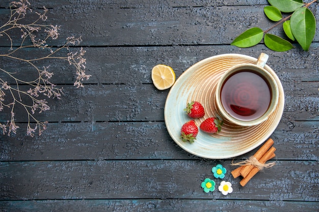 Free photo top view a cup of tea and strawberries on saucer slice of lemon cinnamon flower candies leaves on the dark wooden table