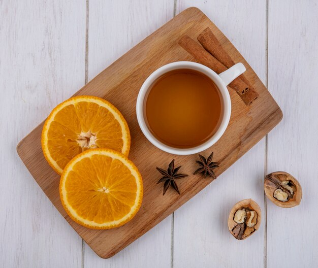 Top view of cup of tea with cinnamon slices of orange on a board with walnuts on a white surface