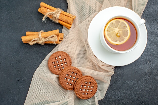 Top view a cup of tea with lemon cinnamon sticks cookies on beige shawl on dark surface