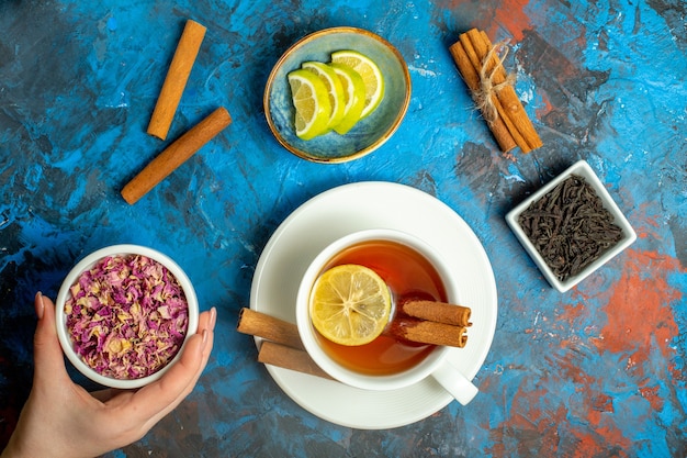 Free photo top view a cup of tea with lemon and cinnamon sticks woman hand holding dried rose petals bowl on blue red surface