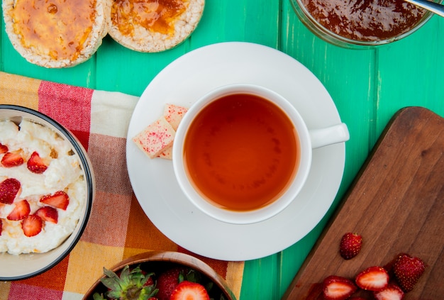 Top view of cup of tea with white chocolate on teabag and bowl of cottage cheese with crispbreads and peach jam on green surface