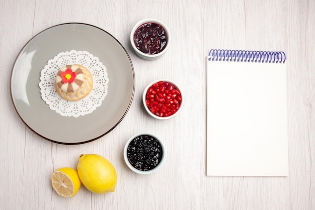 Top view cupcake and jam plate of appetizing cupcake on the lace doily next to the white notebook bowls of seeds of pomegranate jams and lemon on the table