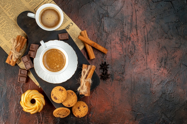 Top view of cups of coffee on wooden cutting board and an old newspaper cookies cinnamon limes chocolate bars on the right side on dark background