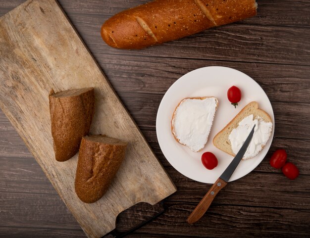 Top view of cut in half baguette and white bread slices with tomatoes and knife in plate on wooden background