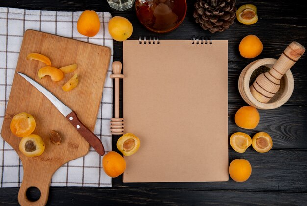 Top view of cut and sliced apricots with knife and note pad with pinecone on wooden background