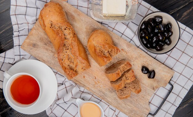 top view of cut and sliced black baguette with cup of tea butter olive on plaid cloth and wooden table