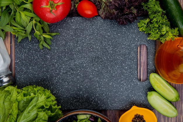 Top view of cut and whole vegetables as tomato basil mint cucumber lettuce coriander with salt black pepper and cutting board on wooden surface