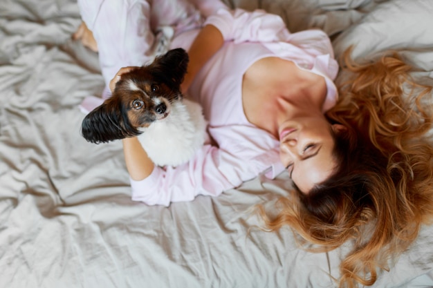 Top view. Cute playful pupppy looking and sitting on bed with pretty ginger girl.