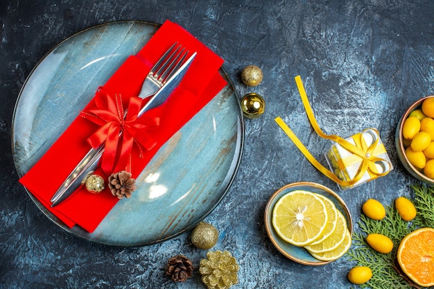 Free photo top view of cutlery set with red ribbon on a decorative napkin on a blue plate and collection of citrus fruits gift accessories conifer cone on dark background