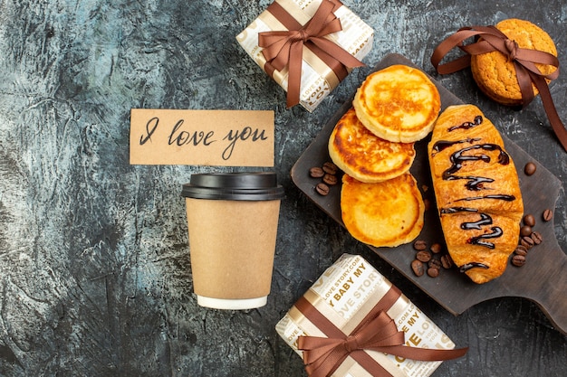 Top view of cutting board with tasty breakfast with pancakes croisasant stacked cookies beautiful gift boxes coffee on dark surface