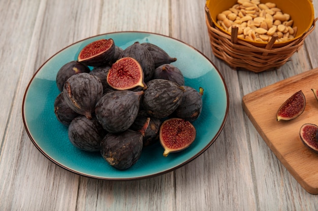 Free photo top view of dark purple mission figs on a blue bowl with slices of black figs on a wooden kitchen board on a grey wooden surface