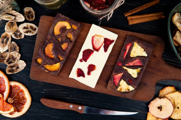 Top view of dark and white chocolate bars on a wooden cutting board with various dried sliced fruits on black background