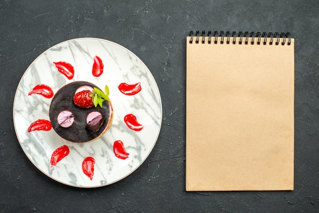 Top view delicious cake with strawberry and chocolate on oval plate a notebook on dark background