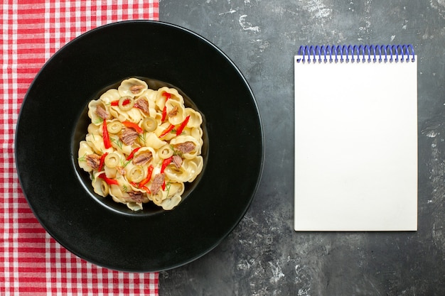 Free photo top view of delicious conchiglie with vegetables and greens on a plate and knife on red stripped towel next to notebook on gray background