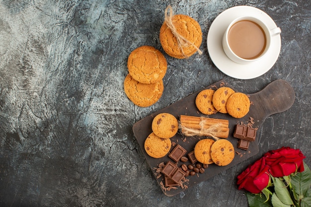 Free photo top view of delicious cookies chocolate bars red roses and a cup of coffee on the left side on icy dark background