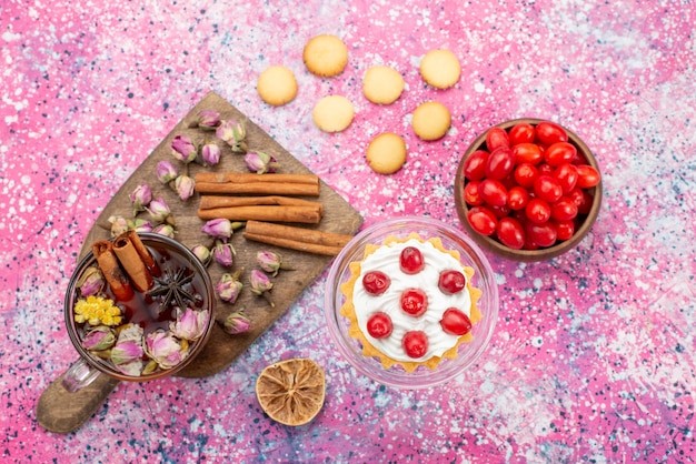 Top view delicious cream cake with fresh red cranberries along with cinnamon cookies and tea on the purple desk sugar sweet