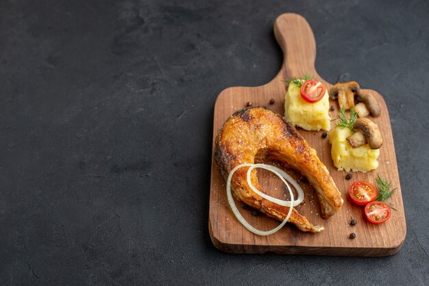 Top view of delicious fried fish meal and mushrooms tomatoes greens on wooden cutting board on the left side on black surface