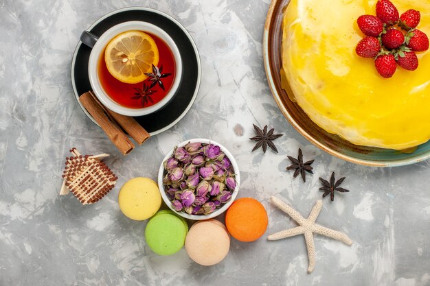 Top view delicious fruity cake with yellow syrup french macarons and cup of tea on white surface cake biscuit sweet sugar bake tea cookie