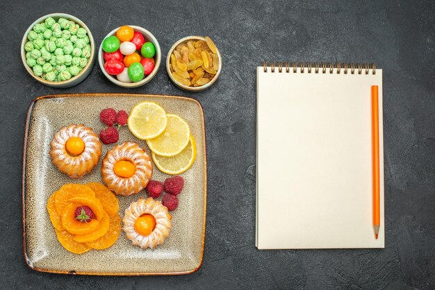 Top view delicious little cakes with lemon slices tangerines and candies on dark background tea fruit biscuit sweet cookies pie