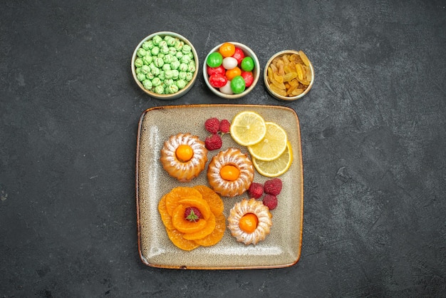 Free photo top view delicious little cakes with lemon slices tangerines and candies on dark background tea fruits biscuit sweet cookie pie