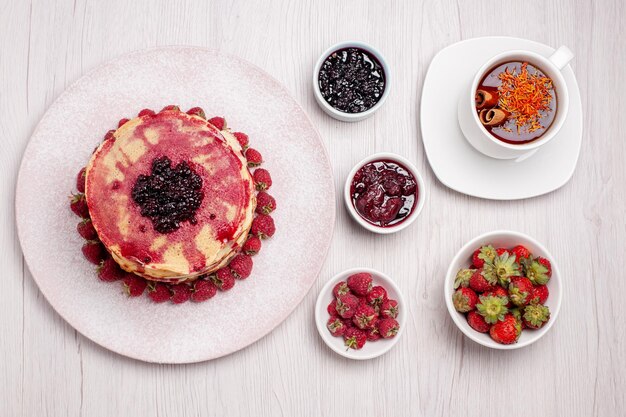 Top view delicious pancakes with strawberries cup of tea on white desk pie biscuit sweet berry fruit cake