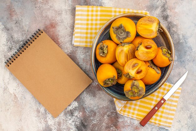 Top view delicious persimmons in a bowl knife yellow kitchen towel a notebook on nude background