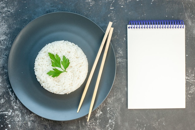 Free photo top view of delicious rice meal served with green and chopsticks on a black plate next to spiral notebook on dark background