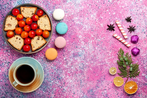 Top view delicious sliced cake with sour fresh plums macarons and cup of tea on the pink surface