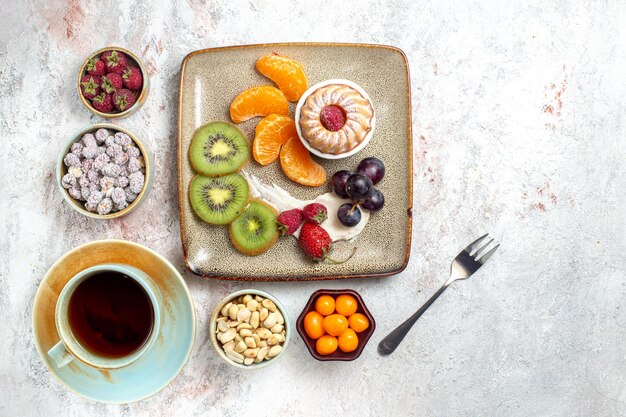 Top view delicious sliced fruits with cake candies and cup of tea on white background fruit fresh tea candy cake biscuit