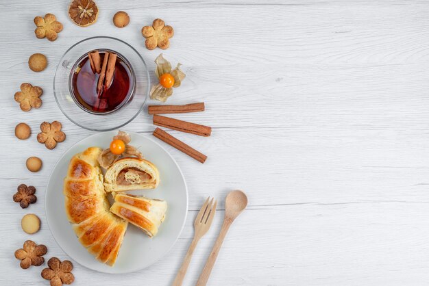 Top view of delicious sliced pastries inside plate with filling along with tea and cookies on white desk, pastry cookie biscuit sweet