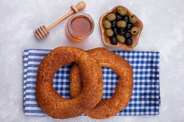 Free photo top view of delicious and soft traditional turkish bagels isolated on a checked cloth with honey on a glass jar and olives on a wooden bowl on a white background