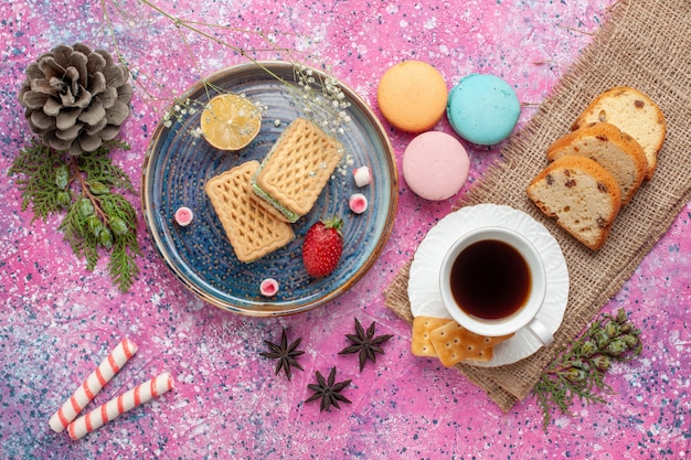 Top view of delicious waffles with french macarons and tea on pink surface