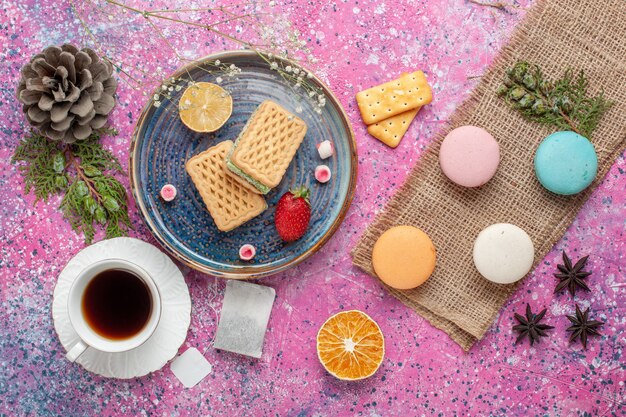 Top view of delicious waffles with macarons and cup of tea on pink surface