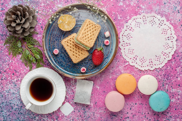 Top view of delicious waffles with yummy french macarons and tea on the pink surface