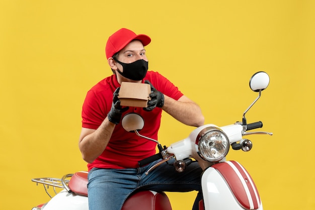 Free photo top view of delivery guy wearing red blouse and hat gloves in medical mask sitting on scooter showing order