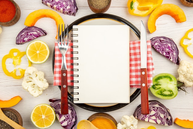 Top view diet written on notepad fork and knife on round plate cut vegetables different spices in bowls on white wooden table