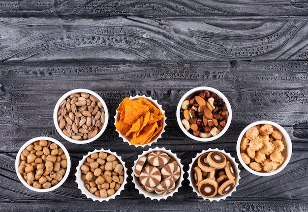 Top view of different kind of snacks as nuts, crackers and cookies in bowls with copy space on dark surface horizontal