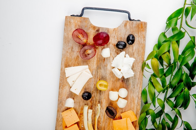 Free photo top view of different kinds of cheese with grape pieces and olives on cutting board on white decorated with leaves with copy space