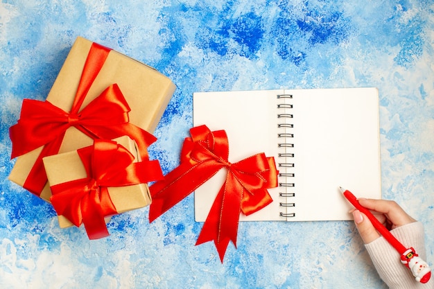 Top view different sizes xmas gifts tied with red bow red bow on notepad pen in female hand on blue table