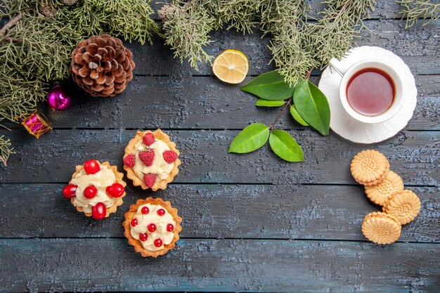 Top view different tarts cone fir-tree leaves christmas toys slice of lemon a cup of tea and biscuits on dark wooden table with copy space