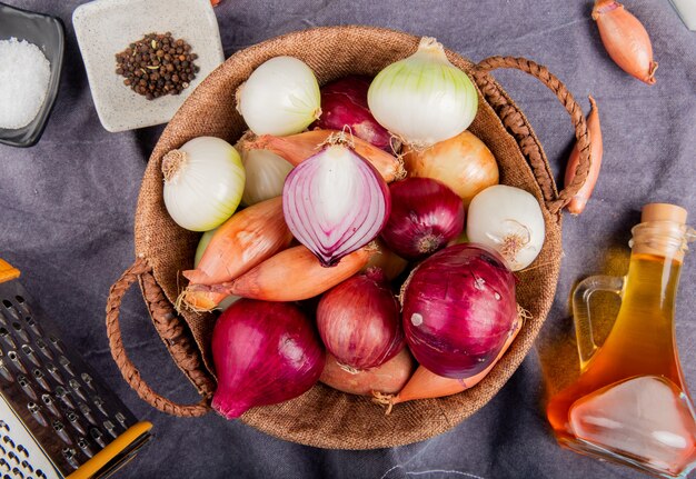 Top view of different types of onion in basket with salt black pepper seeds melted butter grater around on gray cloth