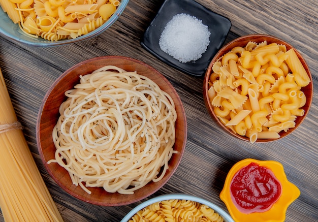 Top view of different types of pasta as spaghetti rotini vermicelli and others with salt and ketchup on wooden surface