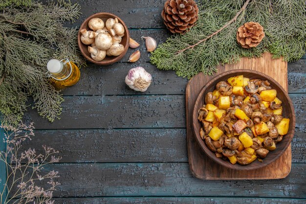 Top view dish and branches dish of mushrooms with potatoes on cutting board next to oil in bottle garlic bowl of mushrooms and branches with cones