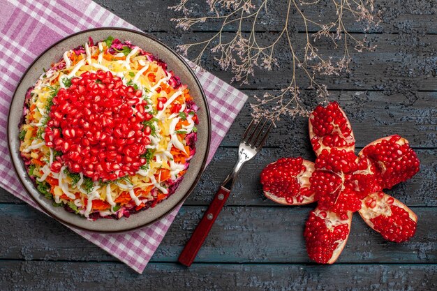 Top view dish and fork pilled pomegranate and dish on the checkered tablecloth next to the branches and fork on the grey table